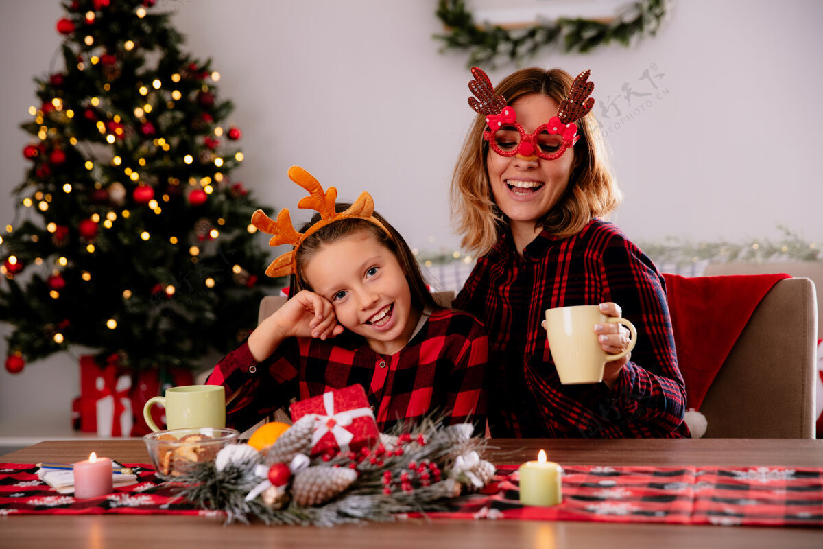 女儿戴着驯鹿眼镜的快乐母亲和坐在餐桌旁的女儿在家里享受圣诞节时光快乐眼镜驯鹿