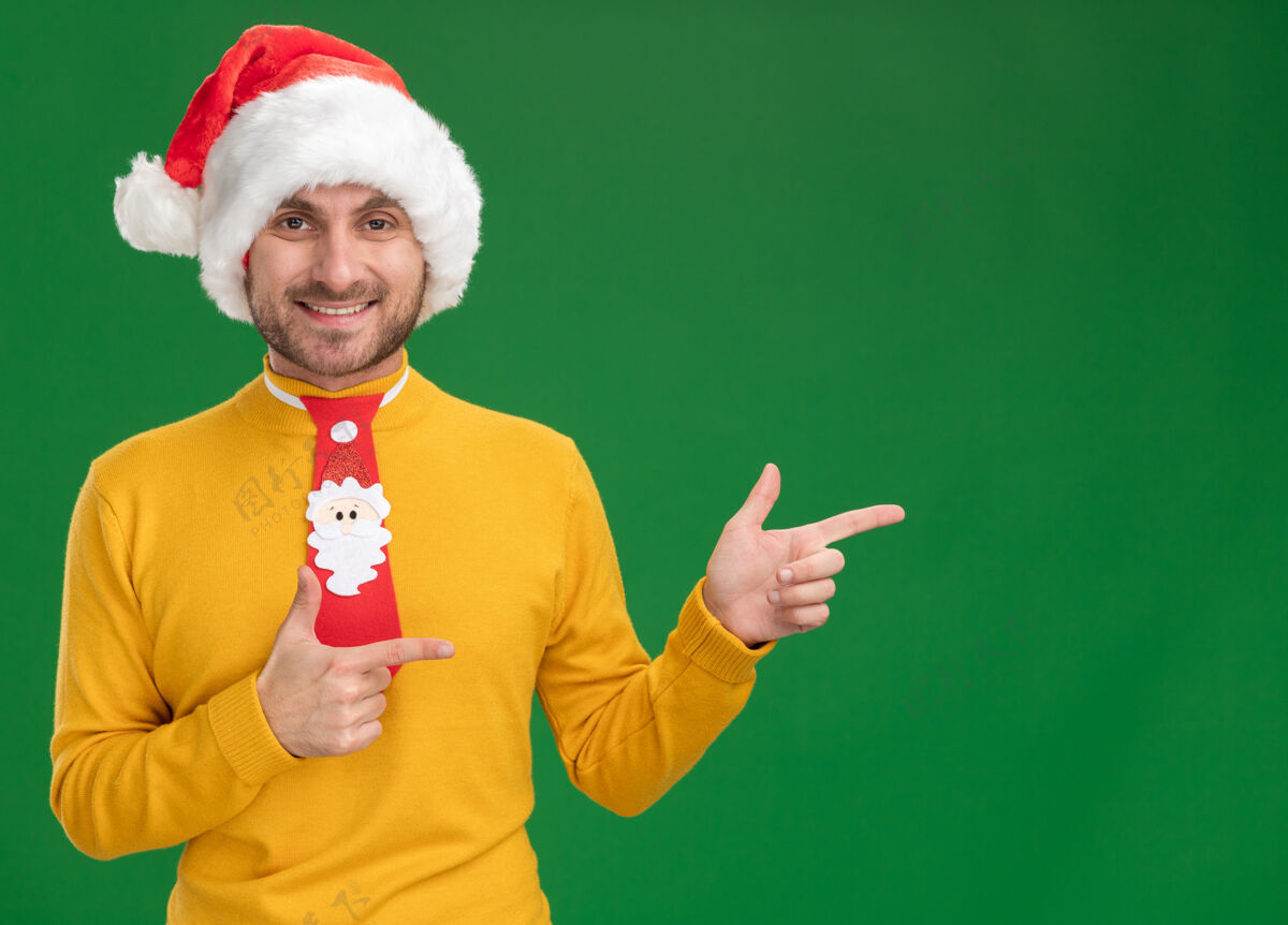 指向戴着圣诞帽打着领带的快乐的白人年轻人看着摄像机指着绿色背景上孤立的一面壁板年轻绿色