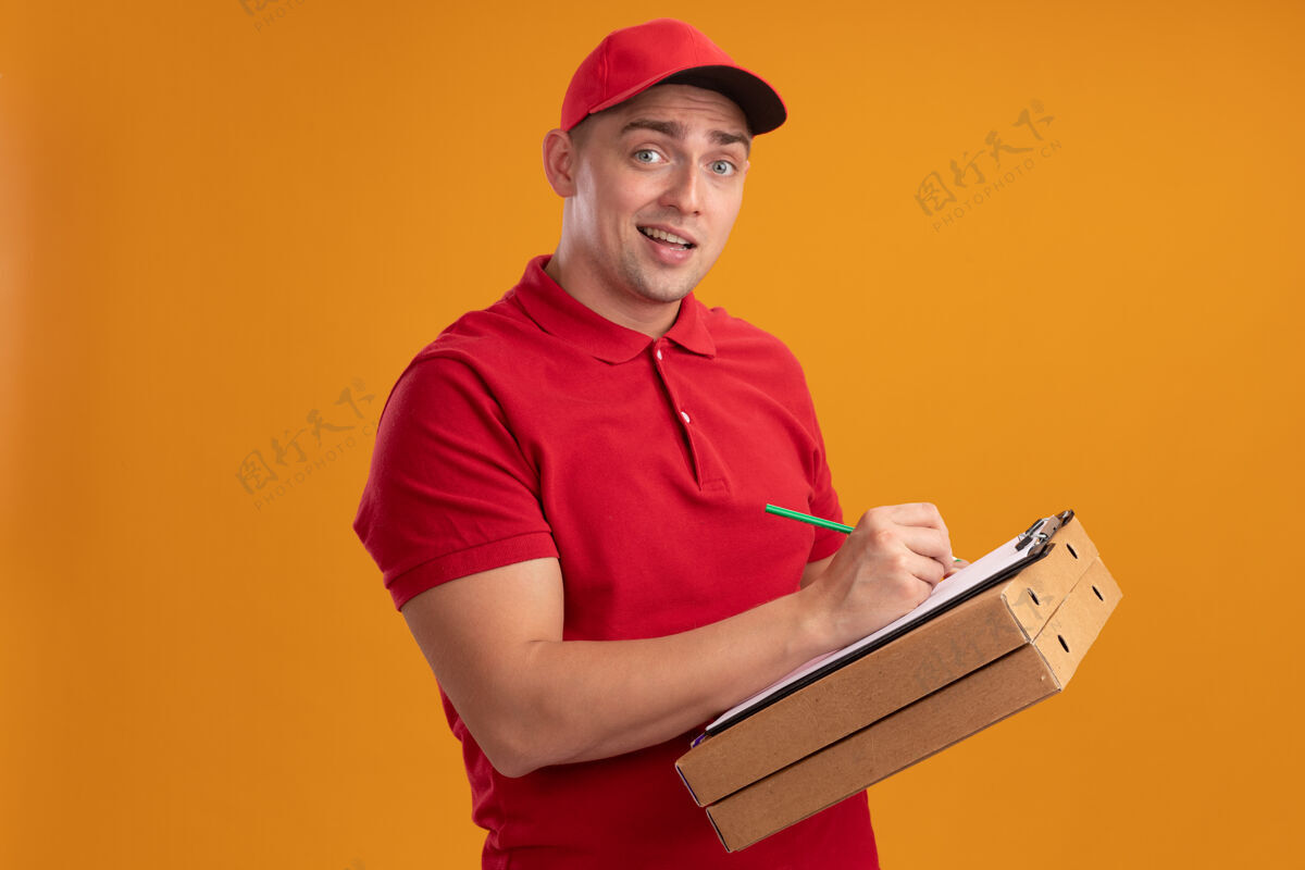 人困惑的年轻送货员穿着制服 戴着帽子 拿着披萨盒 在橙色墙上的剪贴板上写着什么公民披萨什么