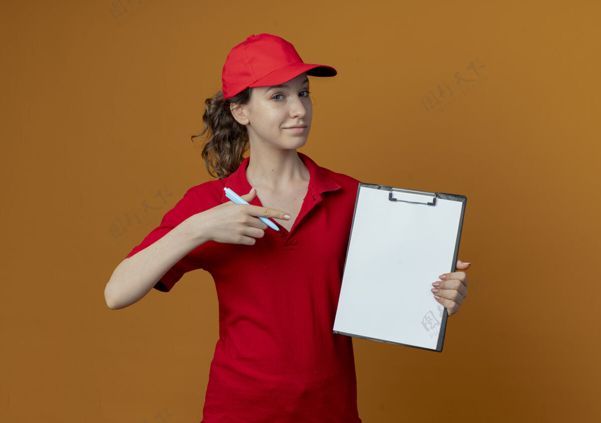帽子自信的年轻漂亮的送货女孩 穿着红色制服 戴着帽子 手里拿着笔和剪贴板 指着橙色背景上孤立的剪贴板 还有复印空间剪贴板制服女孩