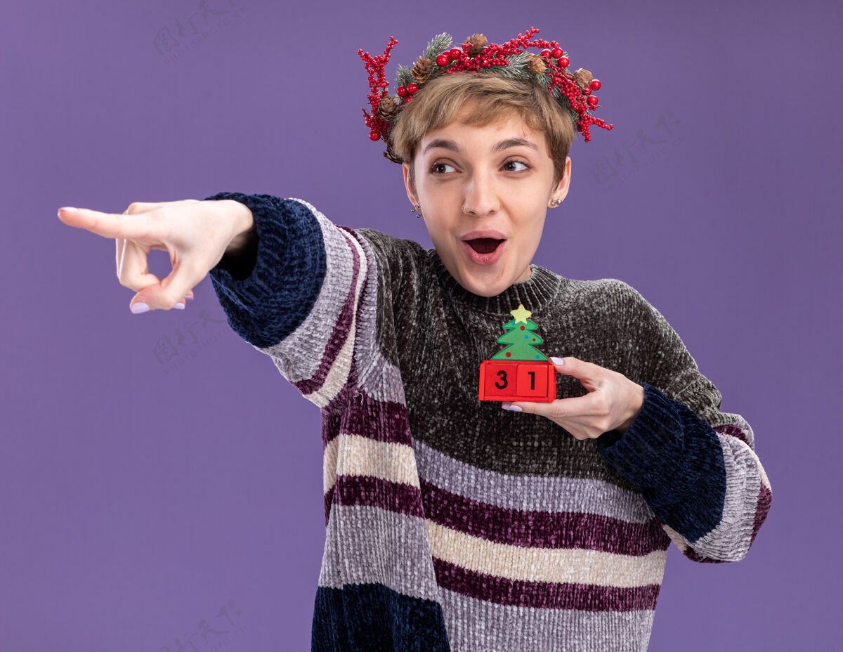 指向令人印象深刻的年轻漂亮女孩戴着圣诞花环手持圣诞树玩具与日期看 并在紫色背景上孤立的一面指着日期圣诞树紫色