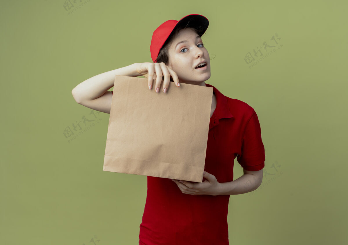 纸张令人印象深刻的年轻漂亮的送货女孩在红色制服和帽子举行纸包看着相机隔离在橄榄绿的背景与复印空间持有漂亮红色