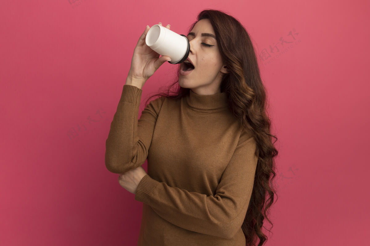 毛衣年轻漂亮的女孩闭着眼睛穿着高领毛衣喝咖啡隔离在粉红色的墙上漂亮脸表情