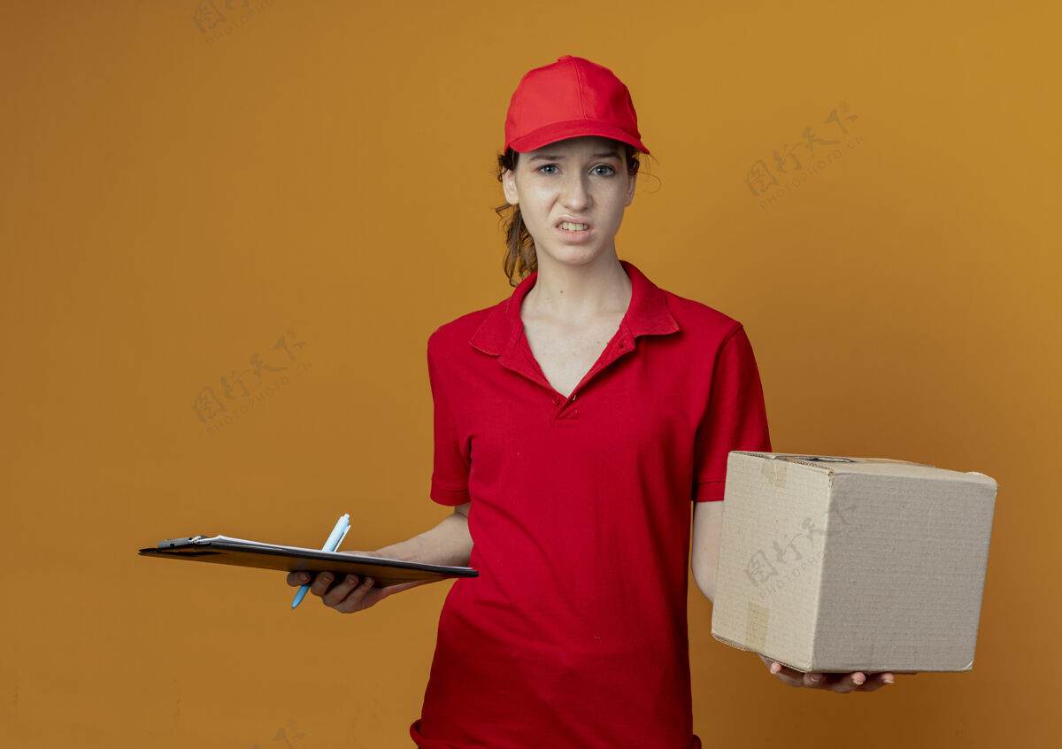 橙色一个年轻漂亮的送货女孩 穿着红色制服 戴着帽子 手里拿着写字板笔和纸箱 孤立地放在橙色的背景上交货背景年轻