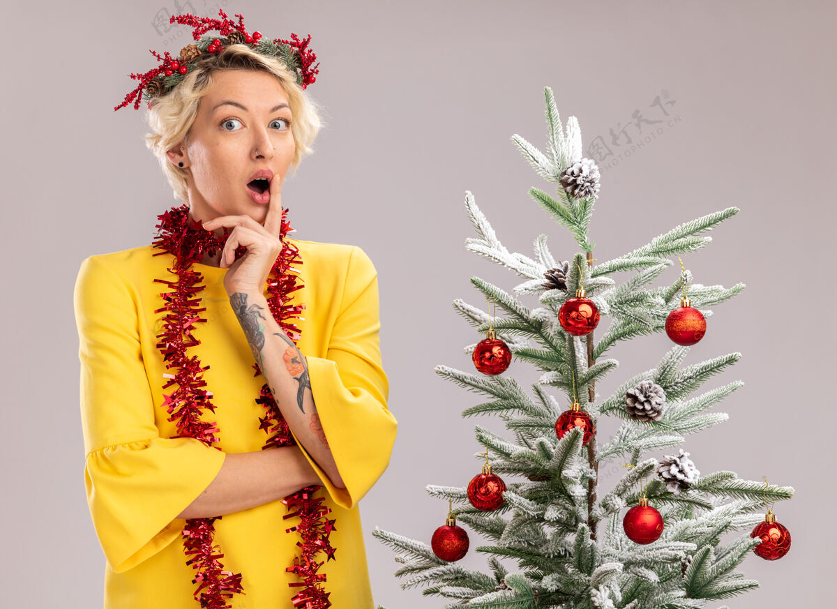 金发令人印象深刻的年轻金发女子头戴圣诞花环 脖子上戴着金属丝花环 站在装饰过的圣诞树旁 看着相机触摸隔离在白色背景上的嘴唇头印象圣诞树
