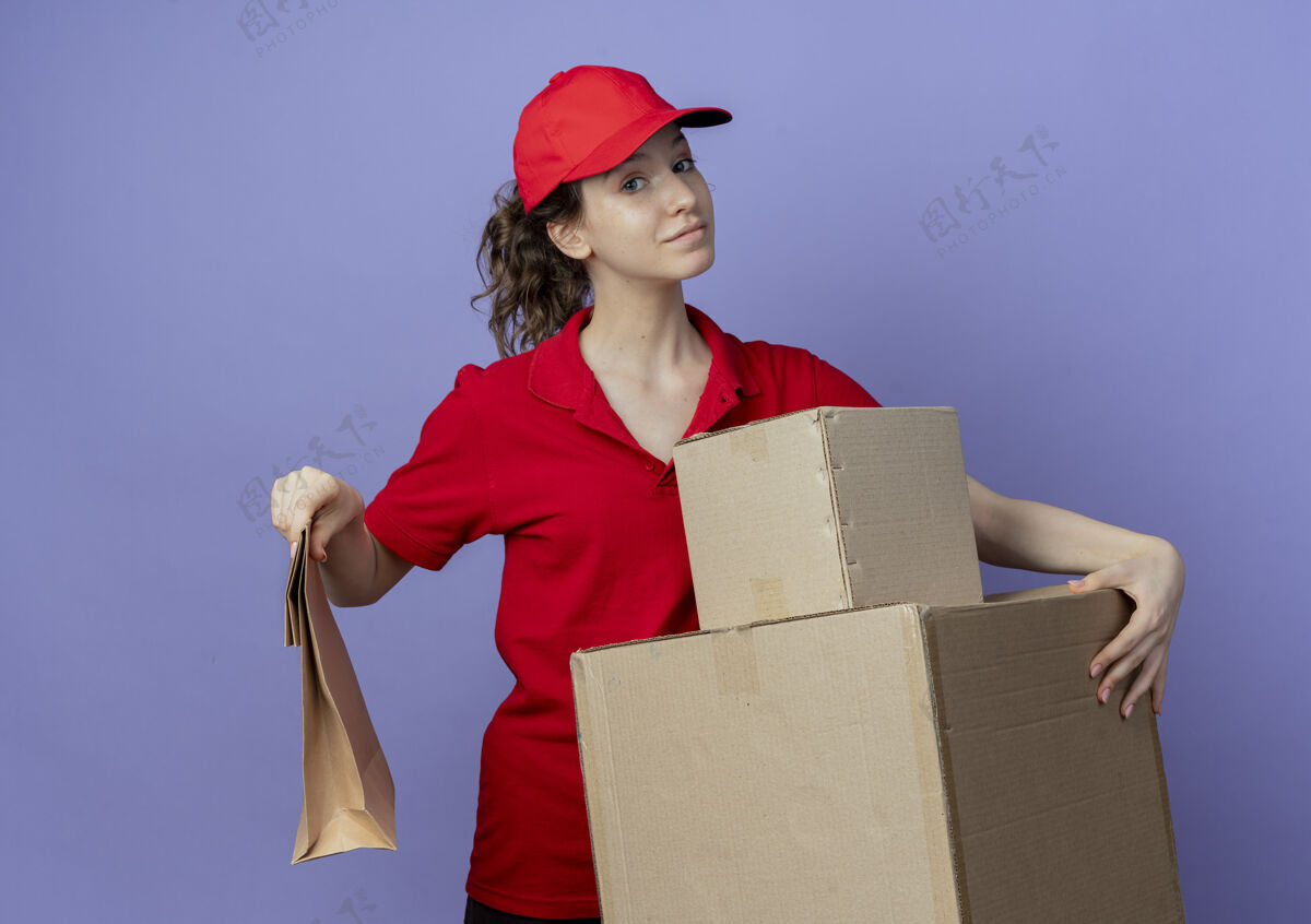 交货高兴的年轻漂亮的送货女孩穿着红色制服和帽子举行纸箱和纸包装隔离在紫色的背景盒子纸年轻