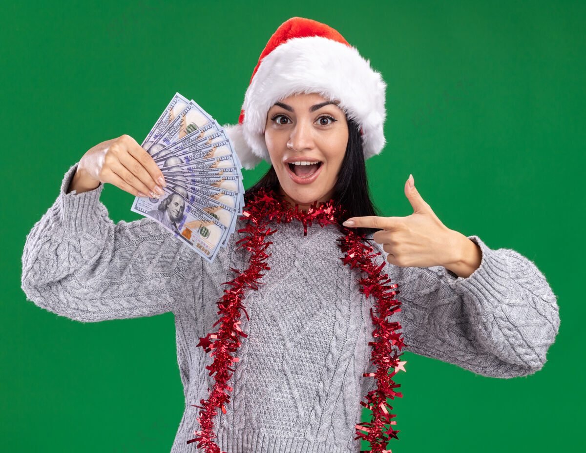 圣诞快乐印象深刻的年轻白人女孩戴着圣诞帽 脖子上戴着金属丝花环 拿着钱 看着隔离在绿色背景上的相机指着女孩绿色