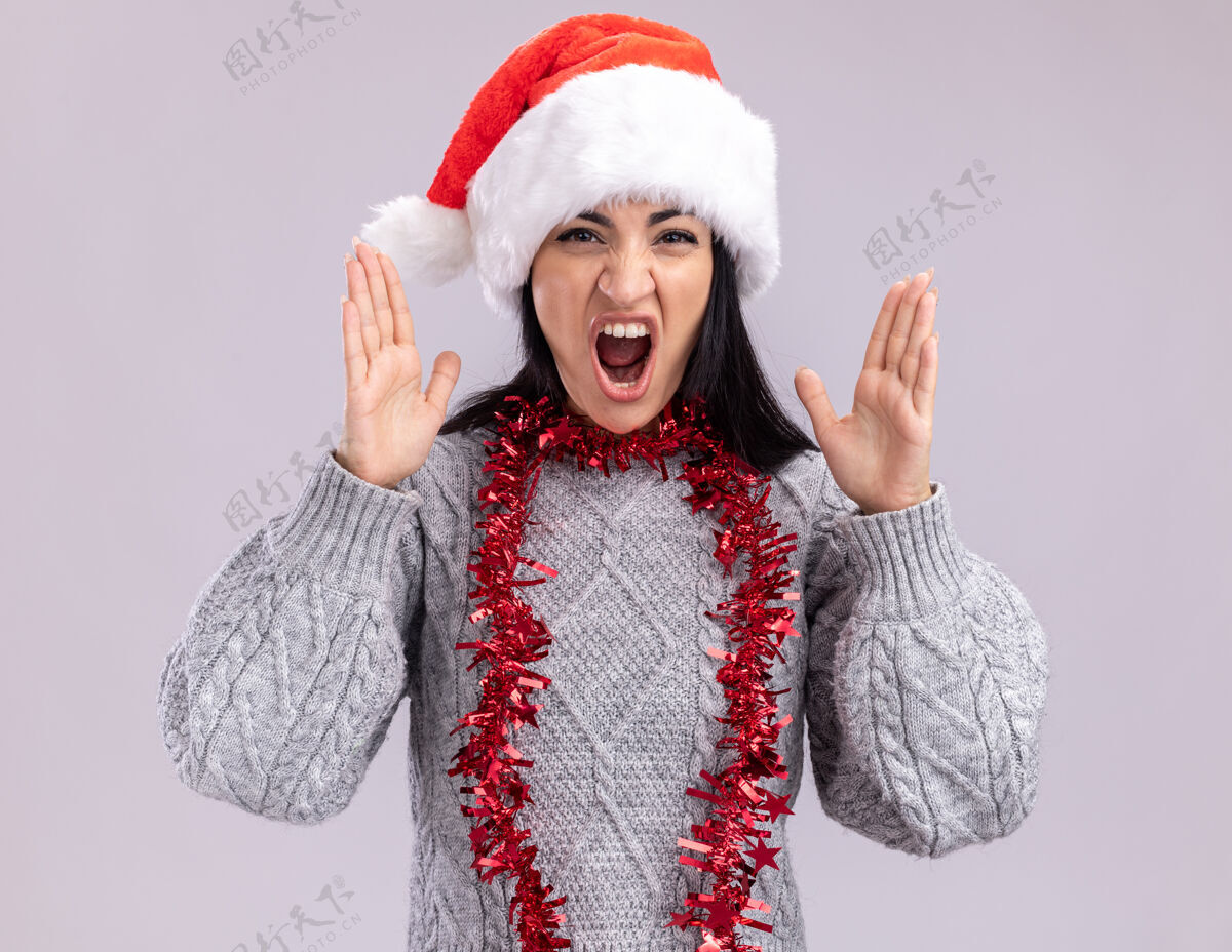 帽子愤怒的年轻白人女孩戴着圣诞帽 脖子上戴着金属丝花环 看着摄像机 双手举在空中 在白色背景上尖叫着孤立花环空气愤怒