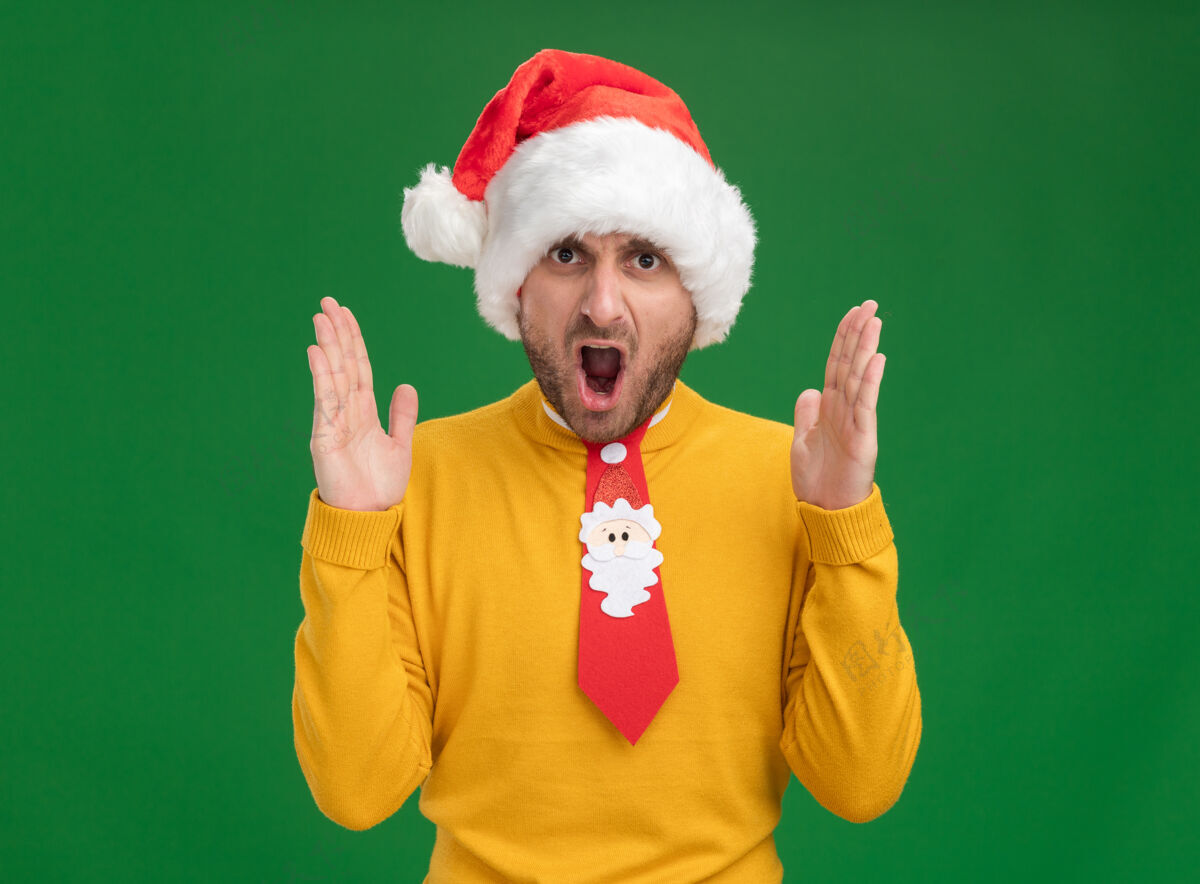 帽子愤怒的白人年轻人戴着圣诞帽 打着领带 看着摄像机 双手举在空中 在绿色背景下尖叫着孤立圣诞男人年轻