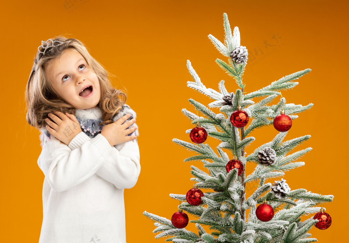 女孩兴奋地抬头仰望站在圣诞树旁的小女孩 戴着头饰 脖子上戴着花环 手放在肩膀上 孤立在橙色背景上肩膀花环脖子