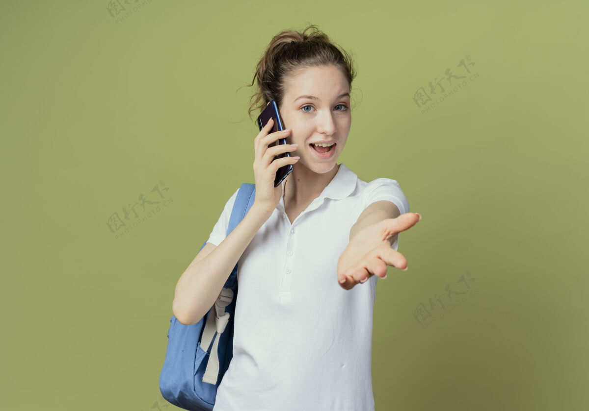 手令人印象深刻的年轻漂亮的女学生穿着背袋讲电话 伸出手对着相机隔离在绿色背景与复制空间伸展印象背部