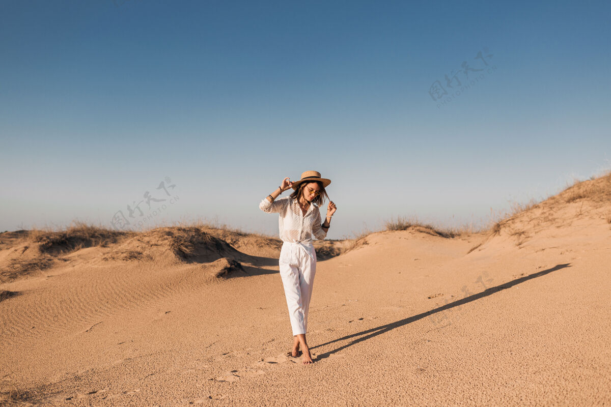 迪拜一个穿着白色衣服 戴着草帽 在沙漠沙滩上行走的时尚美女冒险配件休闲