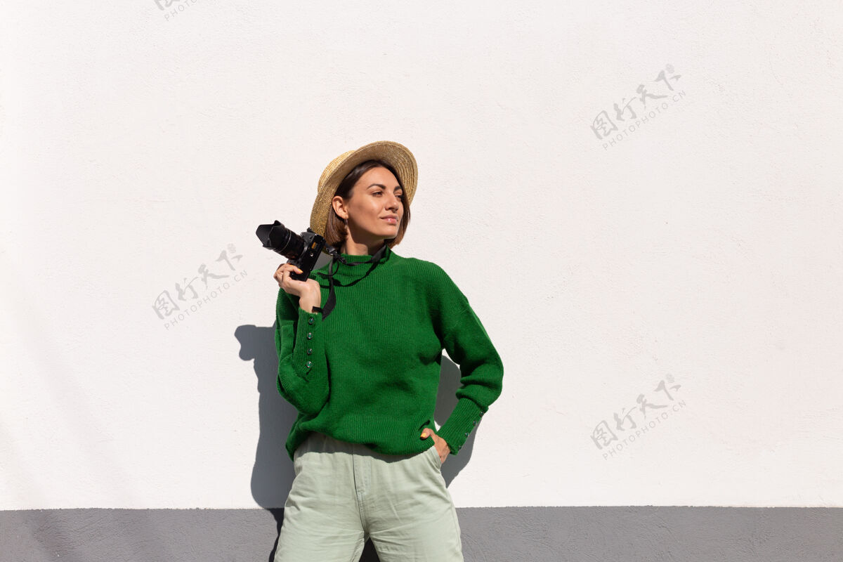 休闲穿着绿色休闲毛衣和帽子的女士户外白墙上快乐的正面游客带着专业相机街道人城市