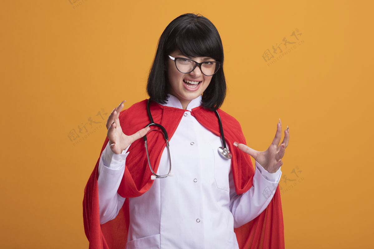 穿兴奋的年轻超级英雄女孩戴着听诊器 穿着医用长袍和斗篷 手上戴着眼镜超级英雄传播听诊器