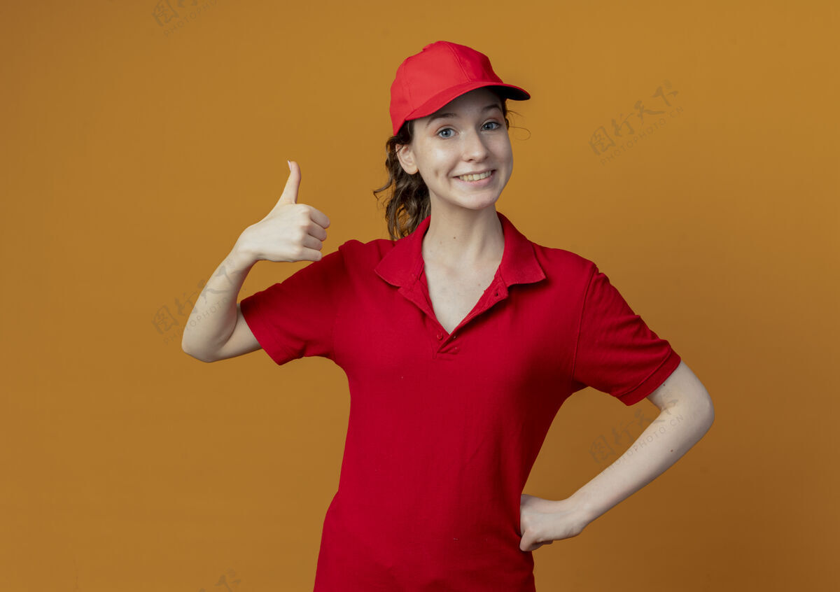 交货微笑着的年轻漂亮的送货女孩 穿着红色制服 戴着帽子 手放在腰上 拇指竖起 孤立地放在橙色背景上 留有复印空间向上女孩腰