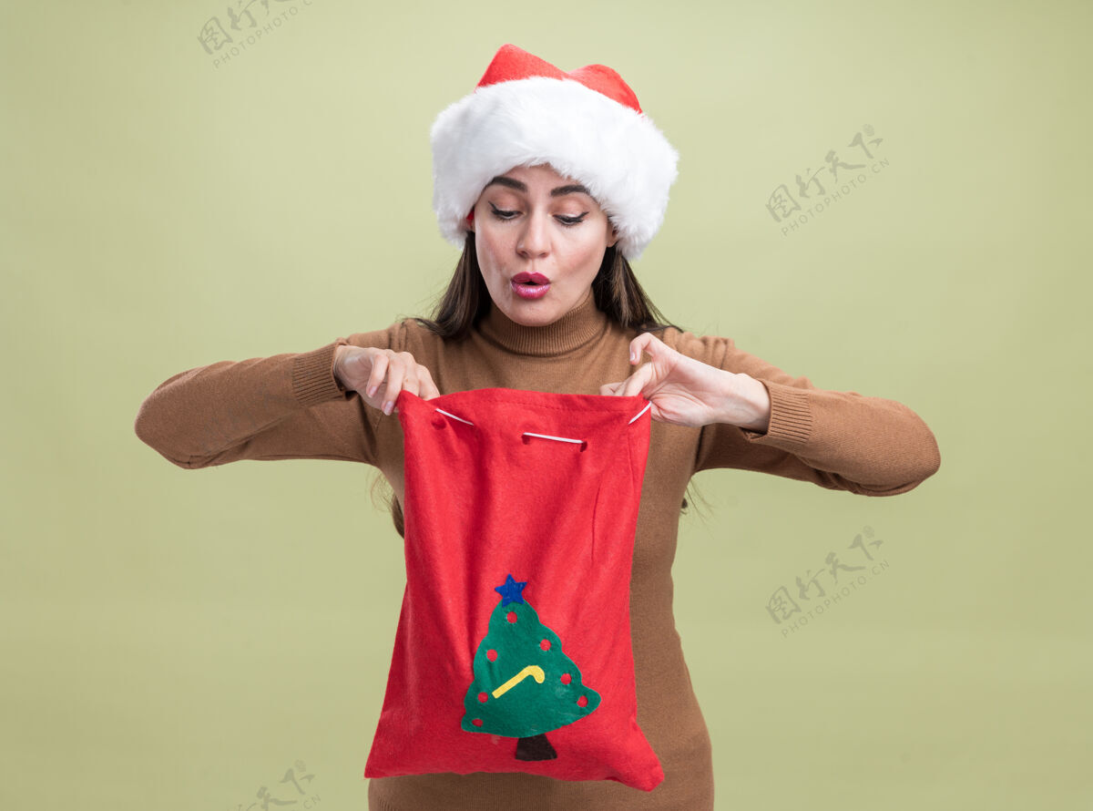 年轻印象深刻的年轻美丽的女孩戴着圣诞帽 抱着圣诞袋看着橄榄绿的背景隔离举行印象绿色