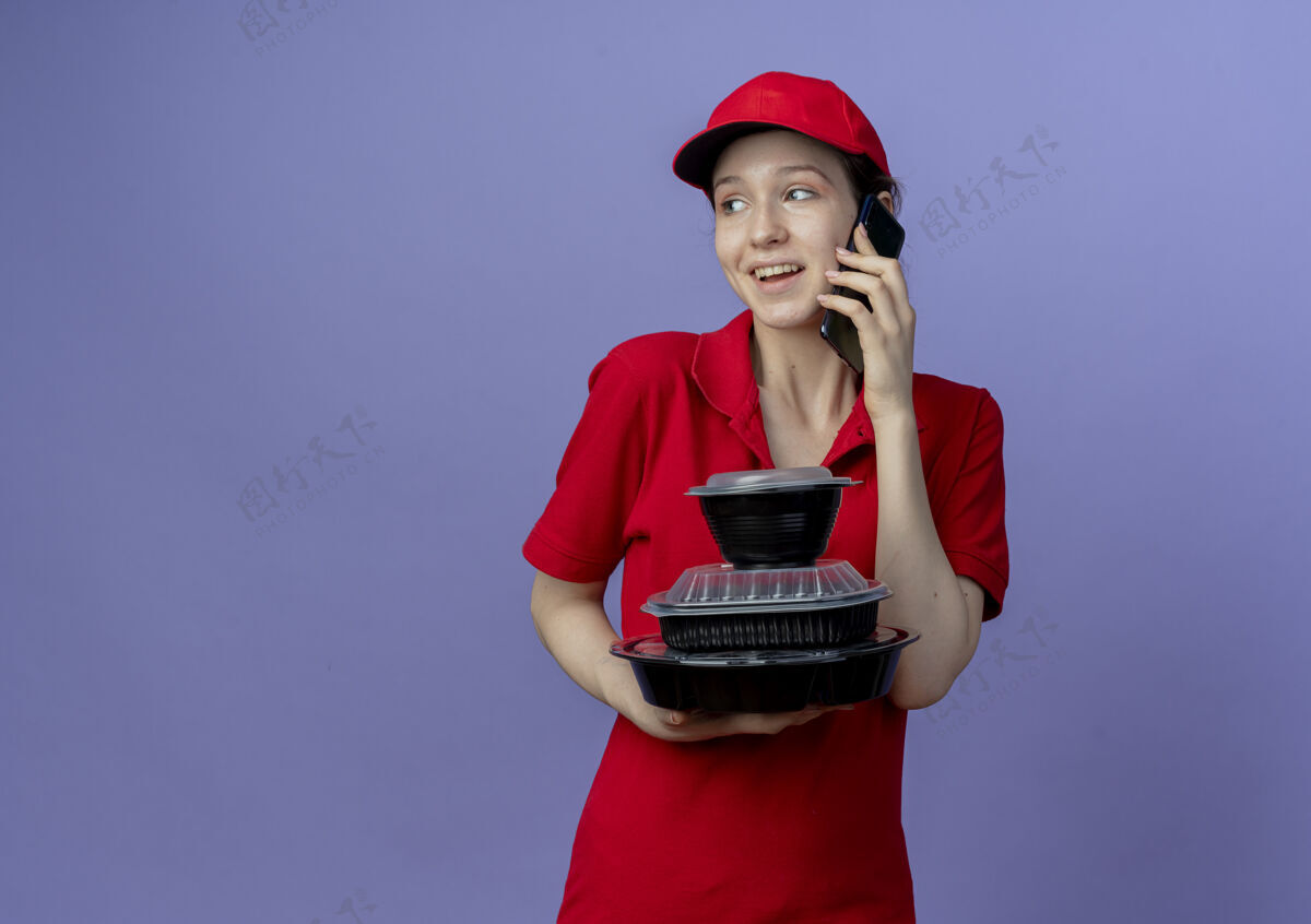 年轻快乐的年轻漂亮的送货女孩 穿着红色制服 戴着帽子 看着一边拿着食物容器 一边在紫色背景上讲着电话 还有复印空间红色制服电话