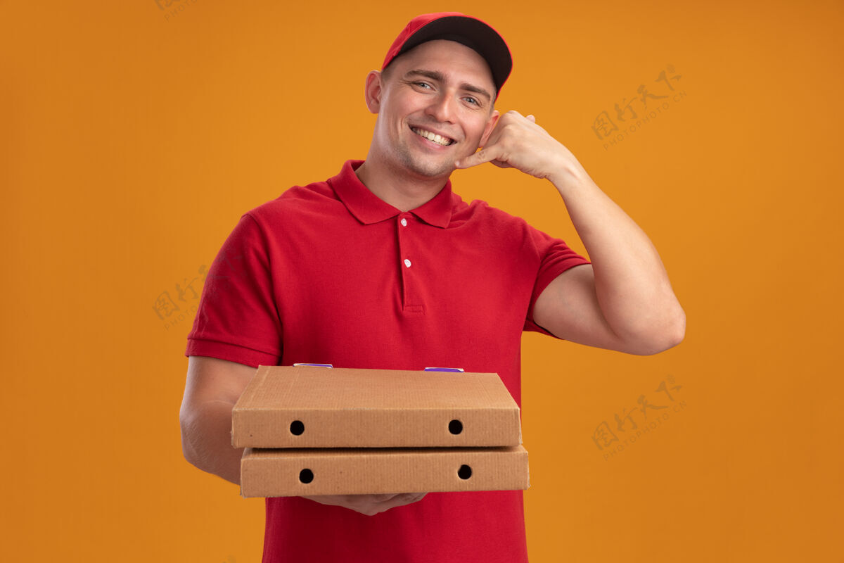 穿着微笑着的年轻送货员身穿制服 戴着帽子 拿着披萨盒 在橙色的墙上显示着电话的手势手势帽子微笑