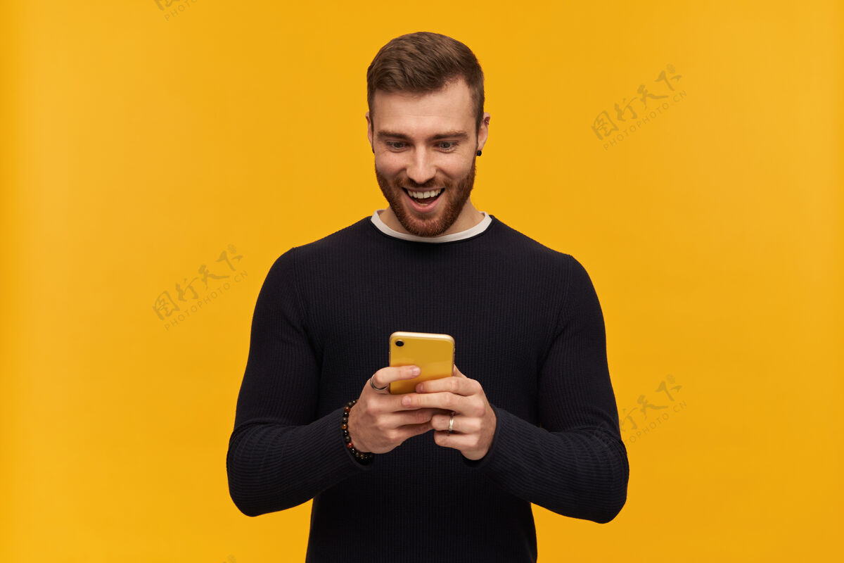 阅读看上去很兴奋的男性 帅哥 深色头发和胡须穿黑色毛衣拿着智能手机看着它阅读信息孤立地站在黄色的墙上帅气模特黑发