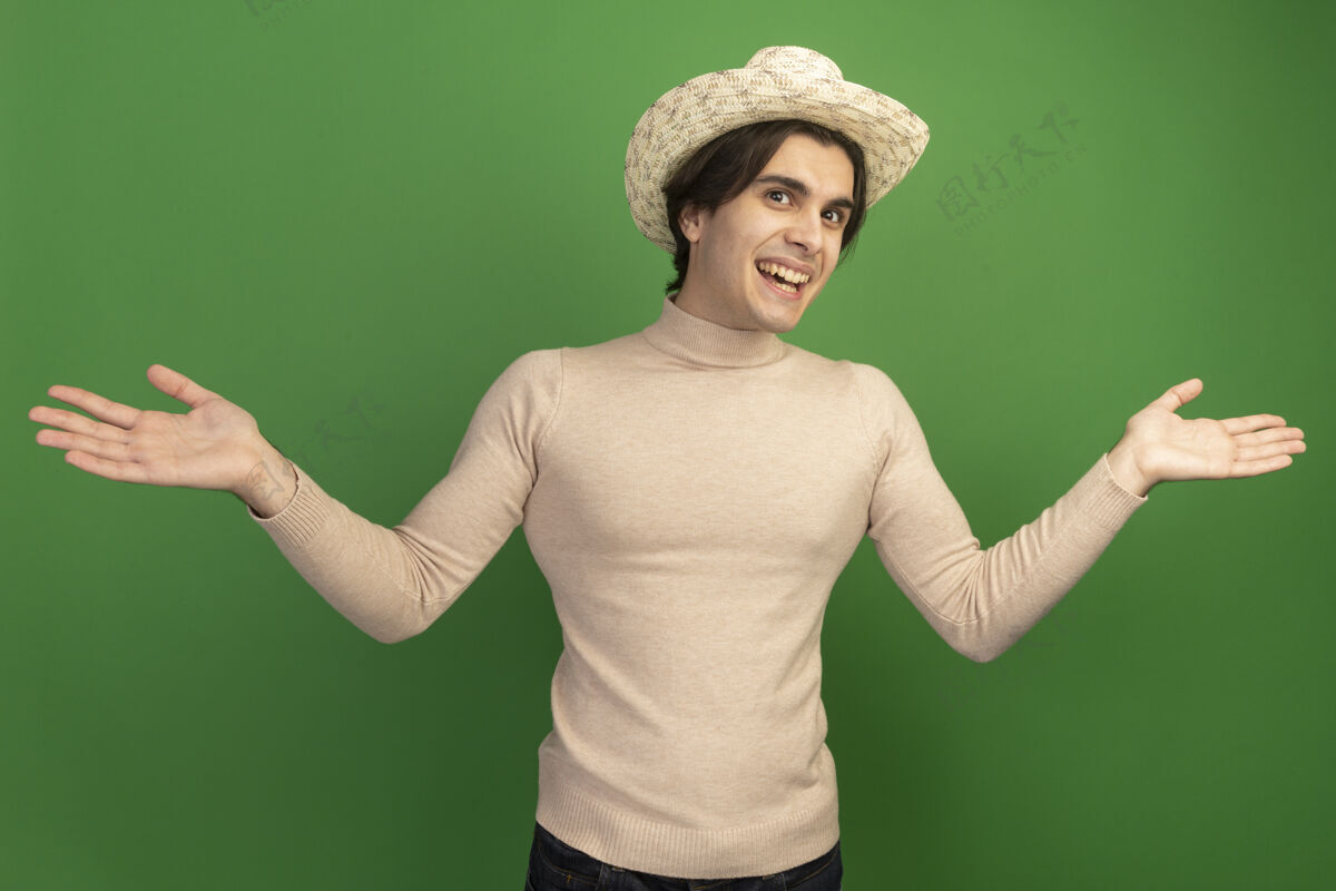 人带着微笑的年轻帅哥戴着帽子摊开双手孤立在绿色的墙上衣服市民帅气