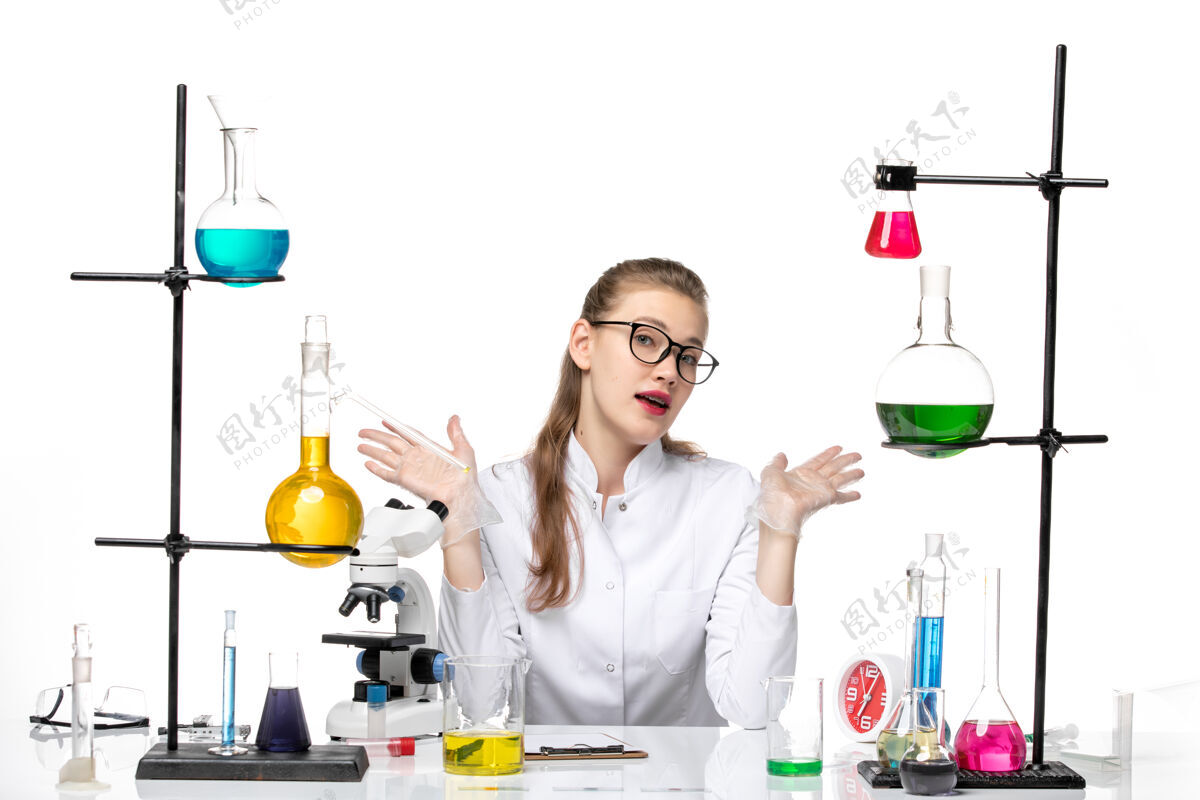 前面前视图穿着白色医疗服的女医生坐在白色办公桌上拿着解决方案病毒化学大流行病毒套装实验室坐着