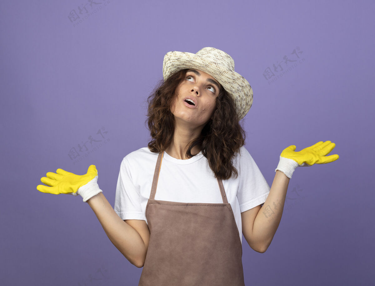 成人惊讶地抬起头来 年轻的女园丁穿着制服 戴着园艺帽和手套 双手摊开向上女性穿