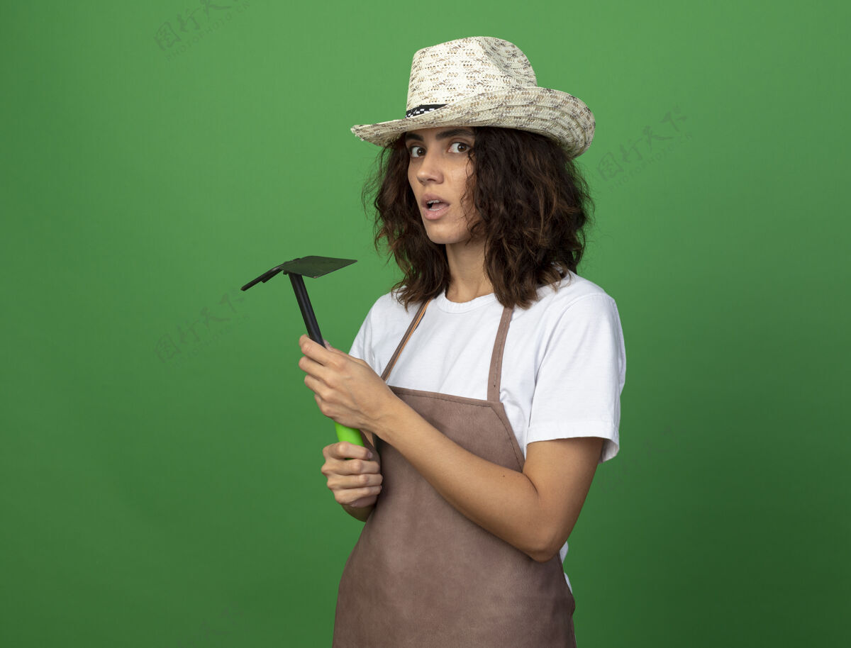 锄头惊讶的年轻女园丁穿着制服戴着园艺帽手持锄头耙子惊喜年轻人女人
