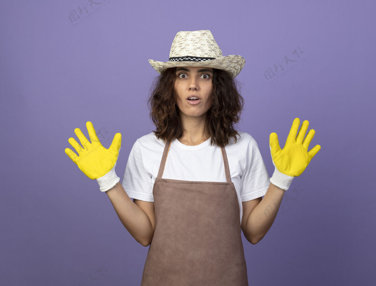 制服穿着制服的年轻女园丁惊讶地戴着园艺帽和手套摊开双手紫色人手