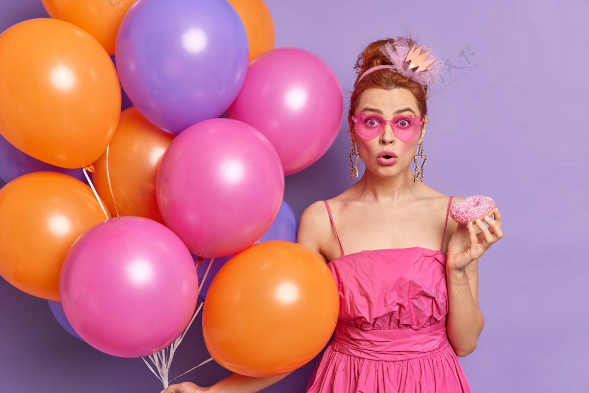 惊喜照片中震惊的红发女子盯着粉红色的阴影 手里拿着美味的玻璃甜甜圈五彩气球发现了令人震惊的消息人印象年轻