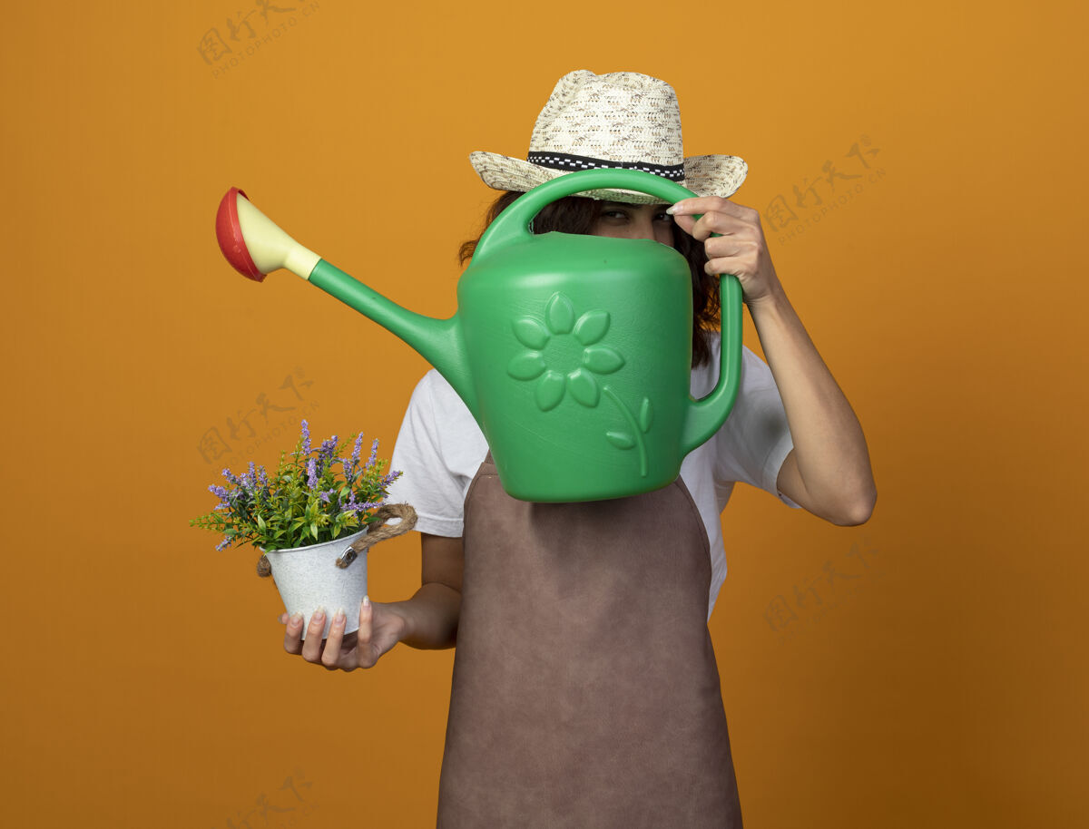 女高兴的年轻女园丁穿着制服 戴着园艺帽 拿着花盆里的花 脸上盖着浇水罐制服浇水园艺