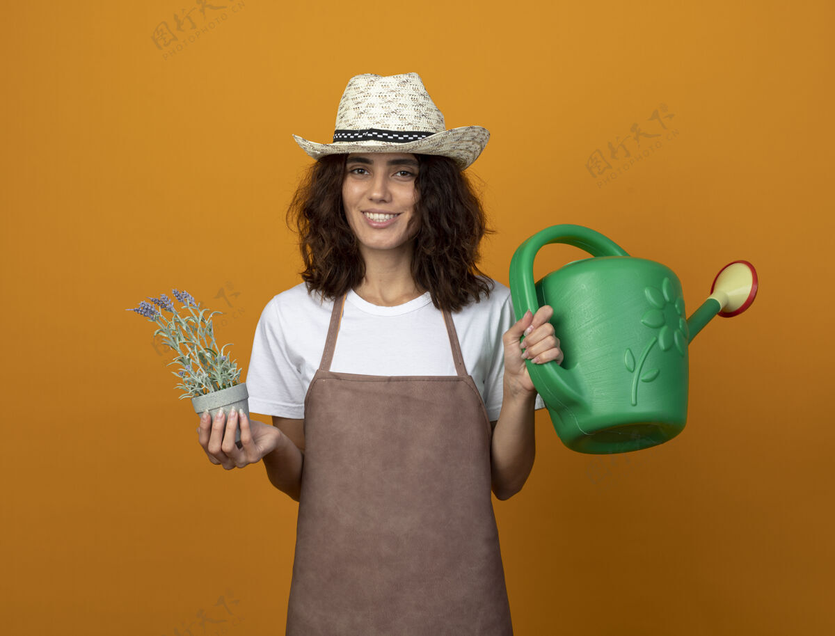 举行微笑着的年轻女园丁 穿着制服 戴着园艺帽 手里拿着花盆里的花和浇水罐制服人女人