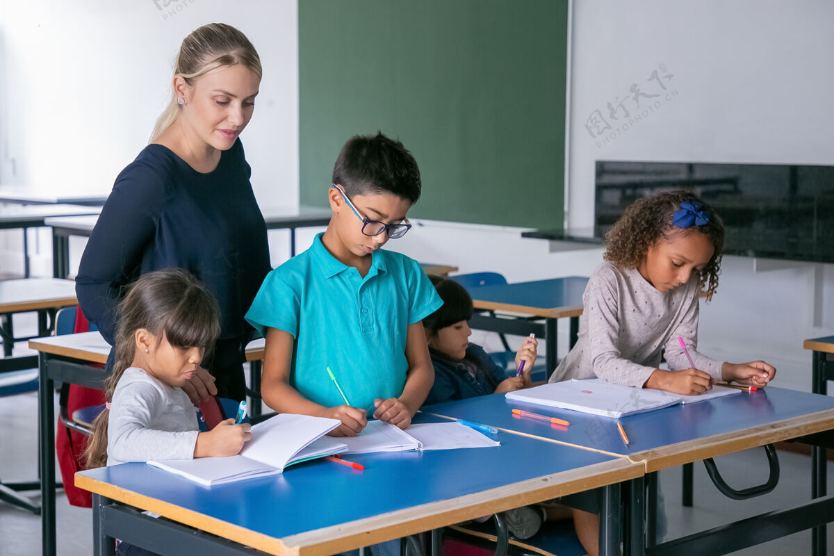 学习积极的女教师看着孩子们在课堂上完成他们的任务 坐在课桌旁 在抄写本上画画和写作班级女人课