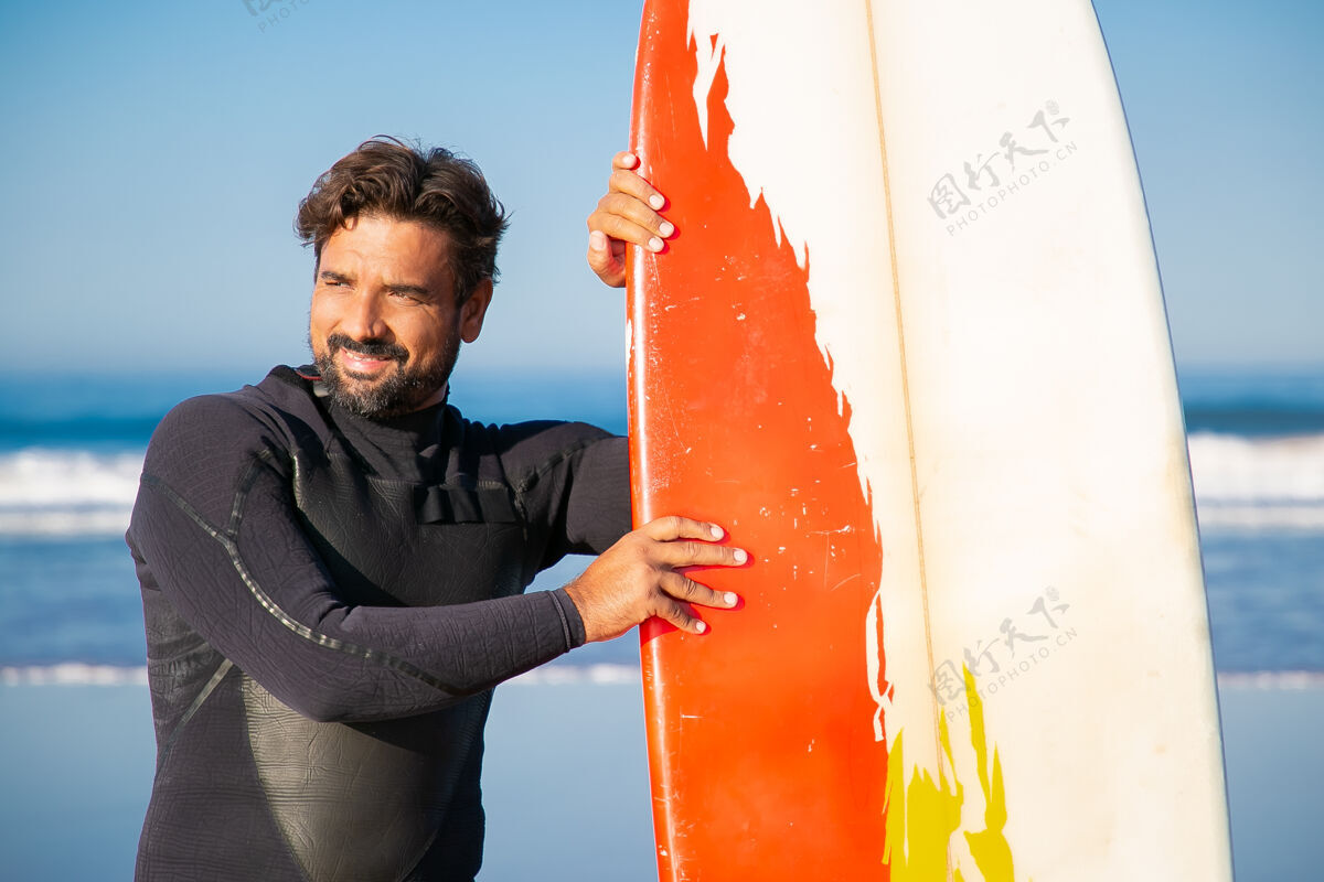 运动员穿着泳衣的快乐男人站在冲浪板旁边 看着远处高加索胡子冲浪者斜靠在冲浪板上微笑着欢呼帅气海洋