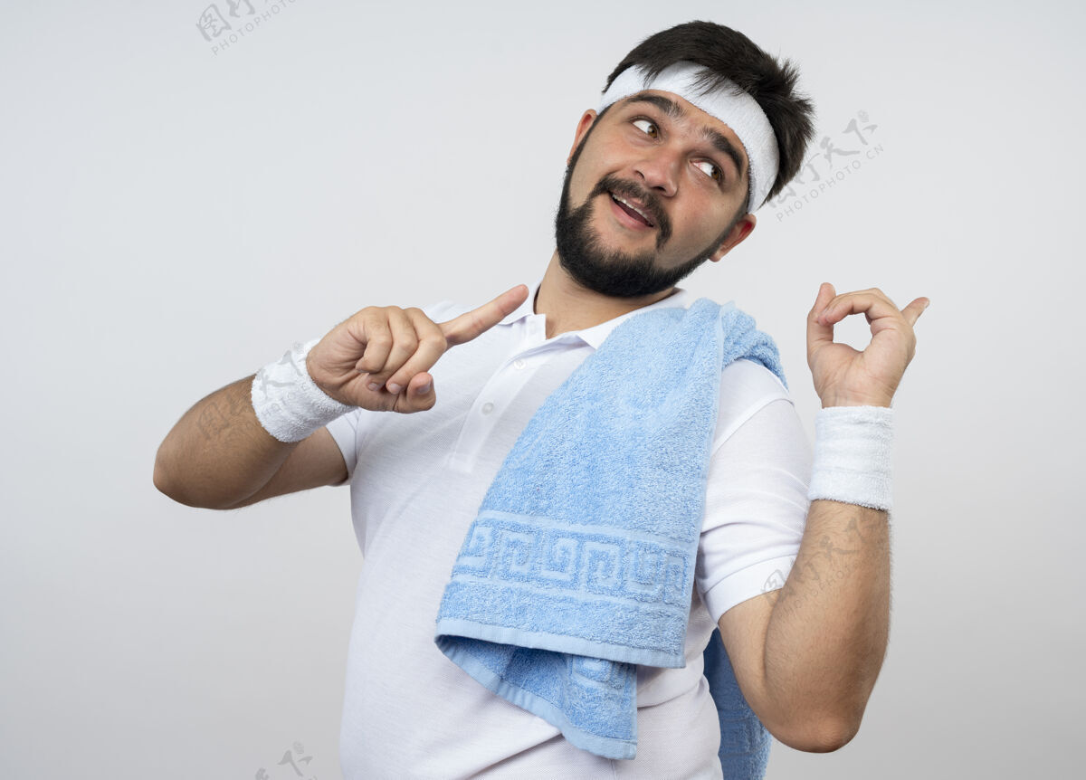 毛巾高兴的年轻运动型男子一边看着戴着头带和手镯 一边把毛巾放在肩上 一边隔离在白色的墙上尖运动请穿