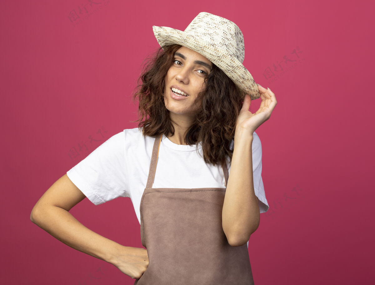 满足微笑着的年轻女园丁 穿着制服 戴着园艺帽 手放在臀部 手拿着粉色隔离帽女人粉红年轻