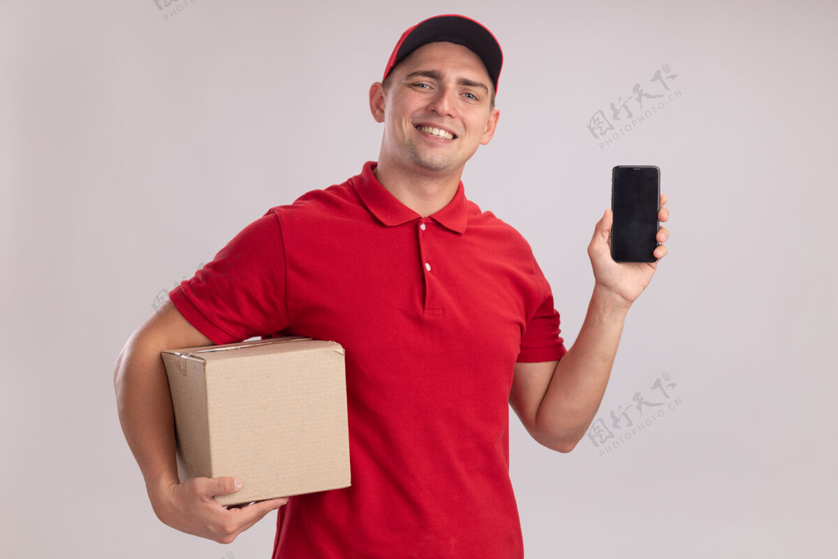 送货微笑的年轻送货员穿着制服 帽子盒和电话被隔离在白色的墙上年轻制服电话