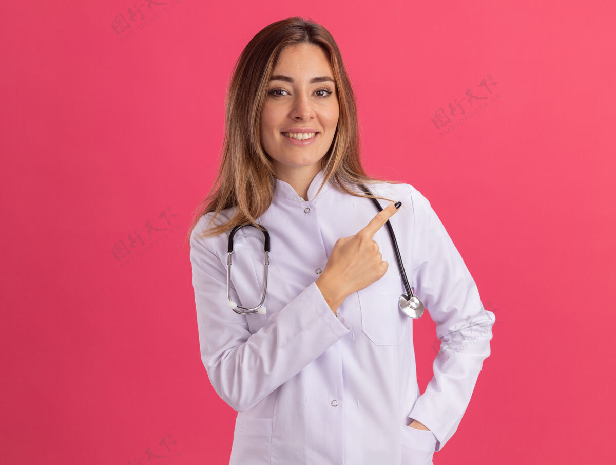 穿面带微笑的年轻女医生穿着医用长袍 听诊器指向隔离在粉红色墙上的一侧 并留有复印空间医疗微笑年轻