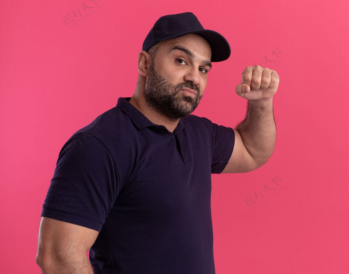 交货自信的中年送货员 穿着制服 戴着帽子 手举拳头 孤零零地站在粉红色的墙上帽子持有拳头