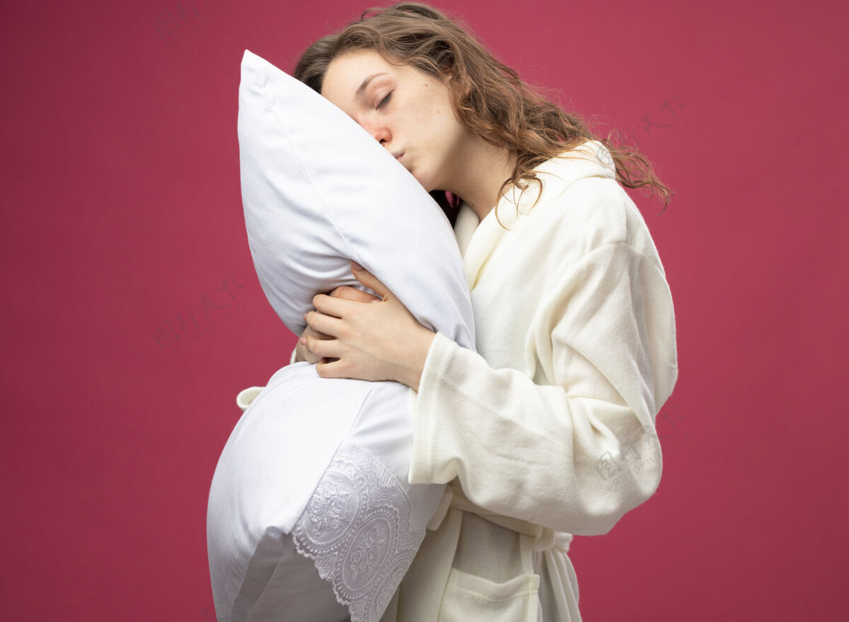 枕头年轻的病女孩闭着眼睛穿着白色长袍抱住枕头孤立在粉红色封闭粉红色长袍