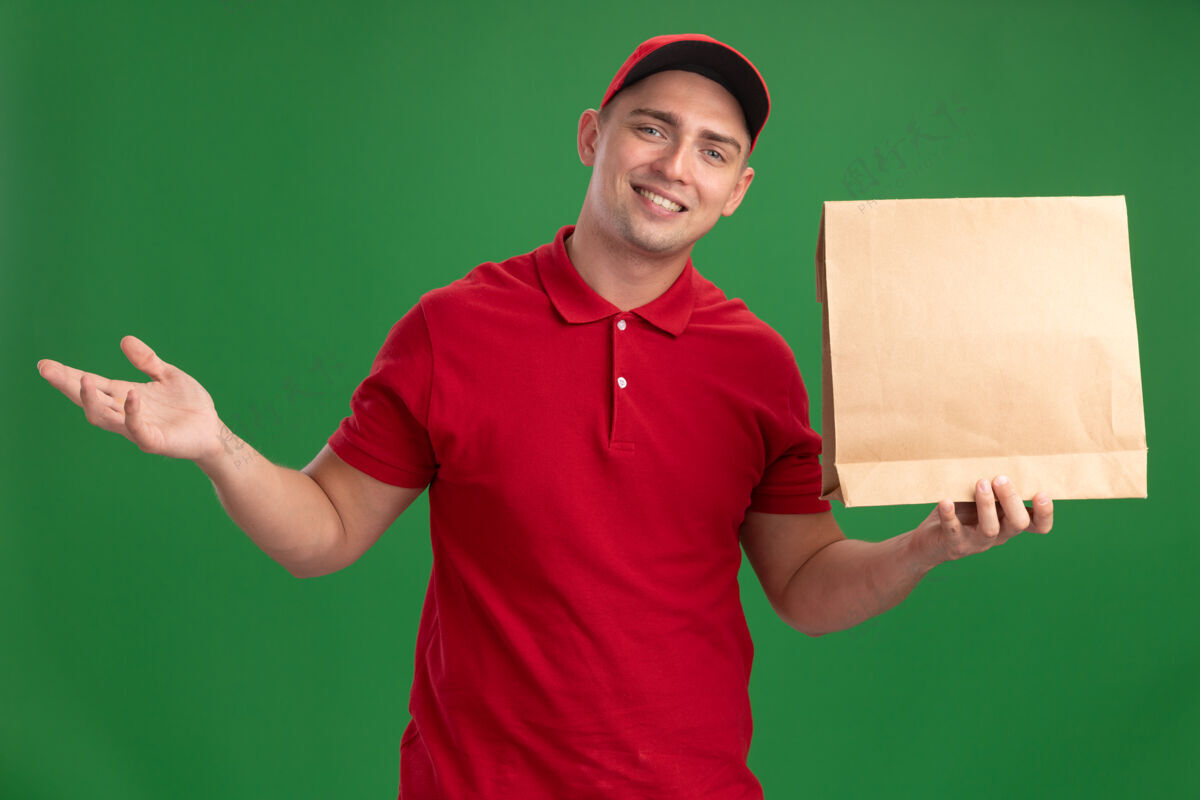 食物面带微笑的年轻送货员穿着制服 戴着帽子 手里拿着纸制食品包 摊开的手被隔离在绿色的墙上年轻人包装拿着