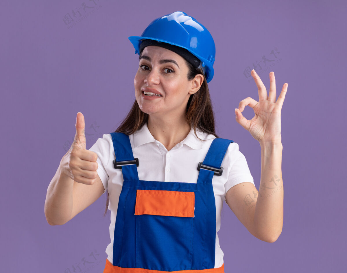 年轻人身着制服的年轻建筑工人微笑着 展示着良好的姿势 在紫色的墙上孤立地竖起大拇指建筑工人制服表演