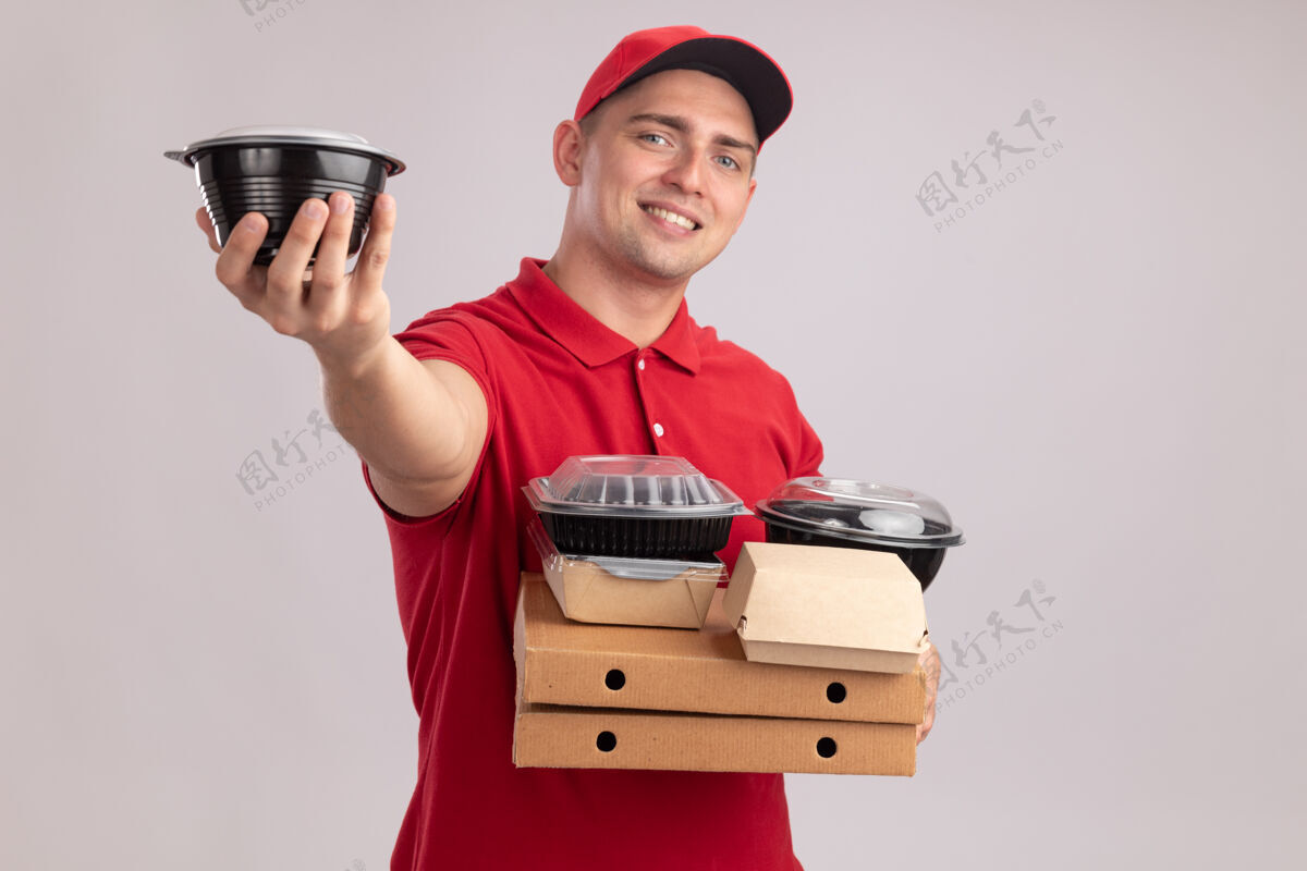 制服微笑着的年轻送货员穿着制服 戴着帽子 拿着比萨饼盒上的食品容器 拿着隔离在白墙上的食品容器食物盒子容器