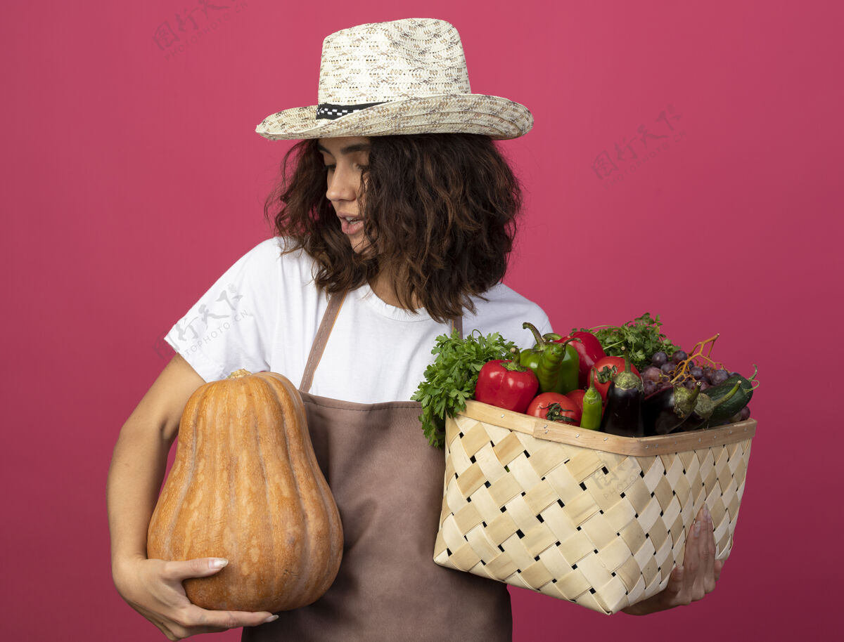 花园高兴的年轻女园丁穿着制服 戴着园艺帽 手里拿着菜篮子 看着手里孤立的南瓜上粉年轻持有园艺