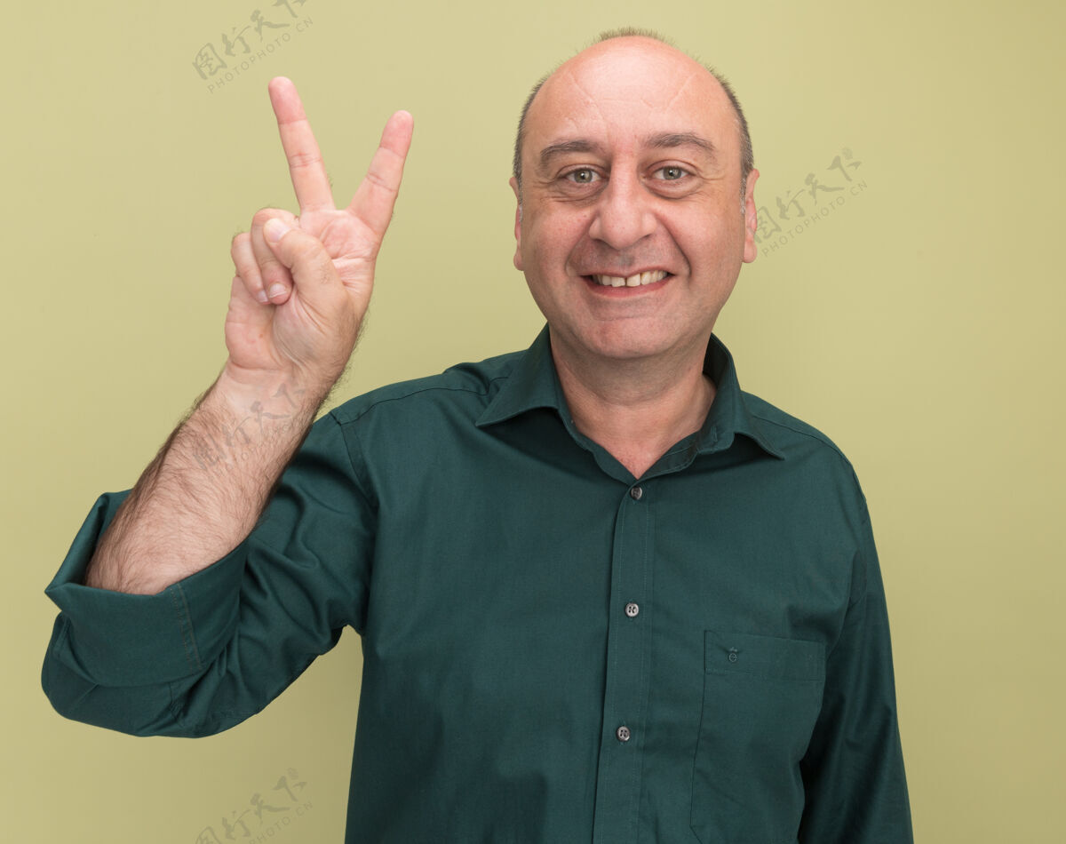 微笑微笑的中年男子身穿绿色t恤 在橄榄绿的墙上显示出与世隔绝的和平姿态中年男人和平