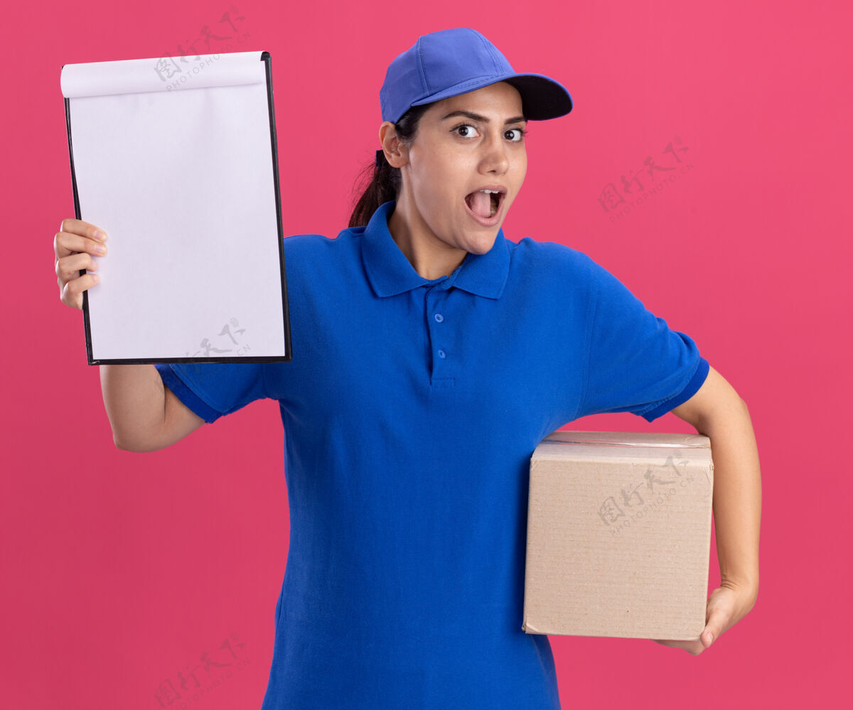 制服惊讶的年轻送货女孩穿着制服 帽子盒和剪贴板隔离在粉红色的墙上穿帽子交货
