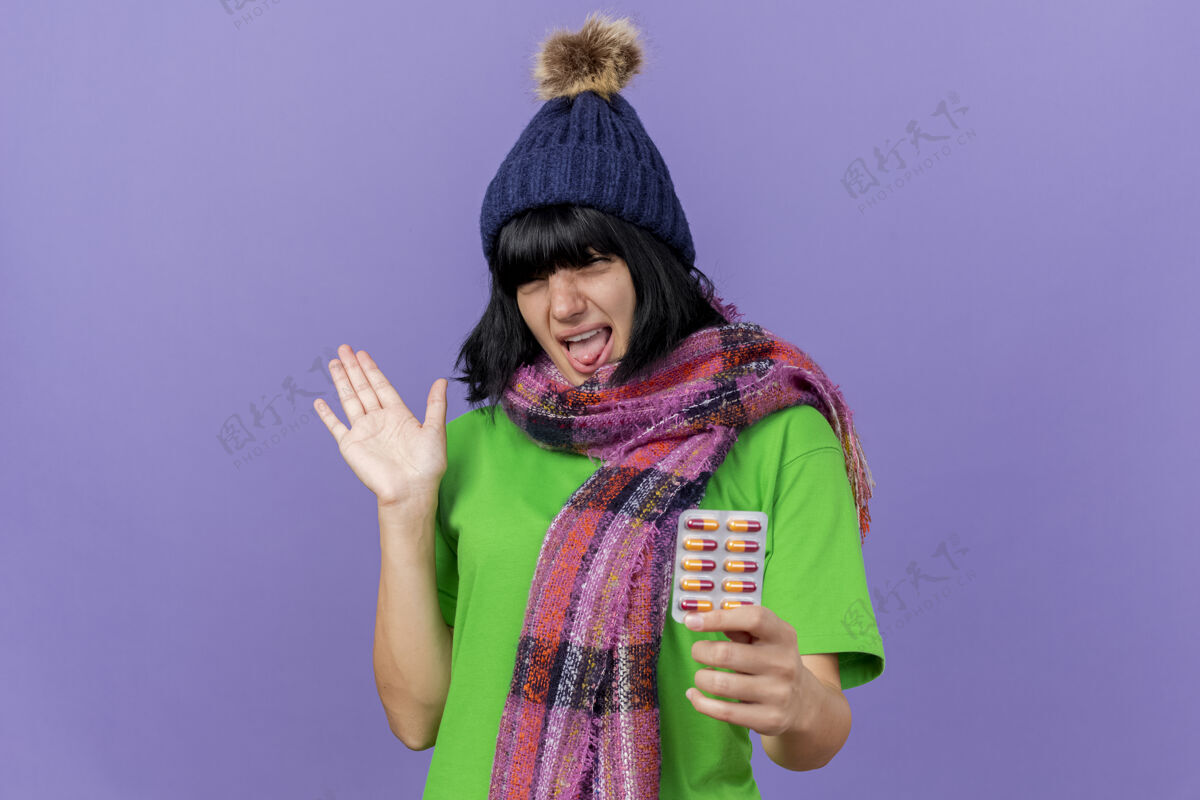 空印象深刻的年轻生病的女人戴着冬天的帽子和围巾 拿着一包胶囊 看着前面显示空的手孤立在紫色的墙壁上复制空间疾病表演表情