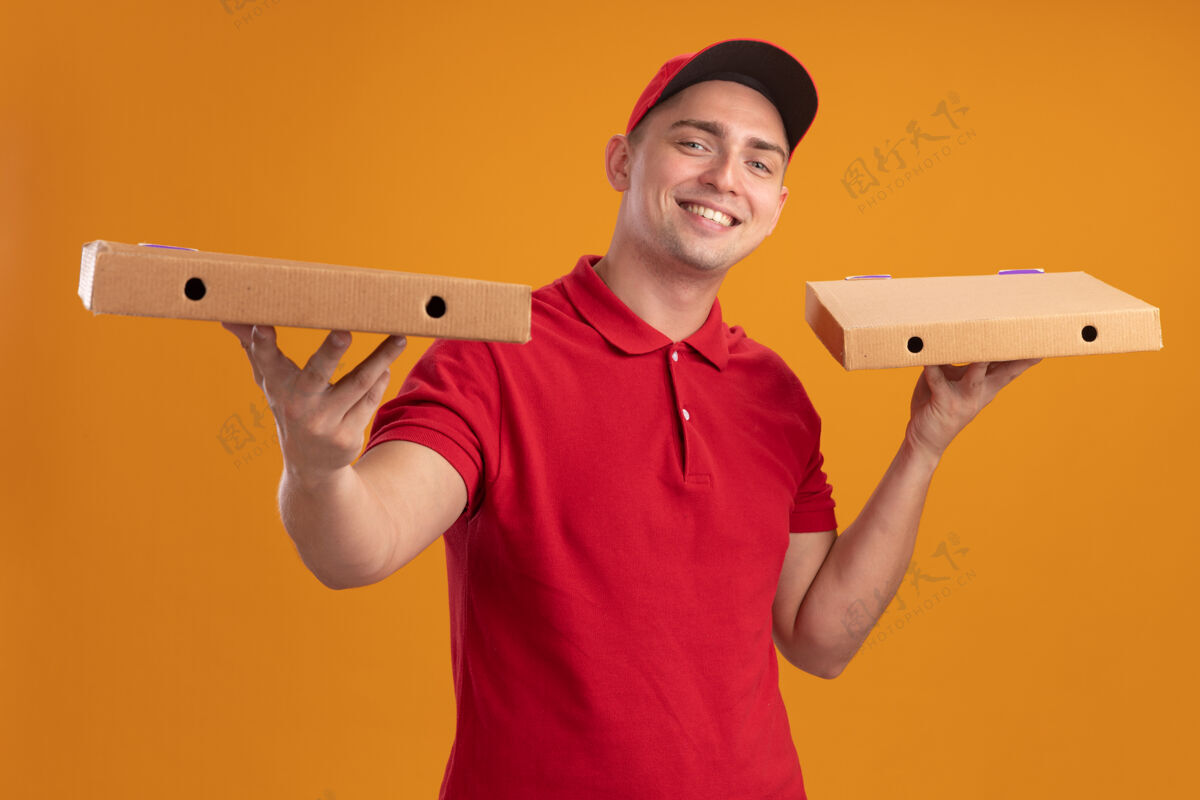 年轻人微笑着的年轻送货员穿着制服 戴着帽子 拿着隔离在橙色墙上的比萨饼盒帽子拿着披萨