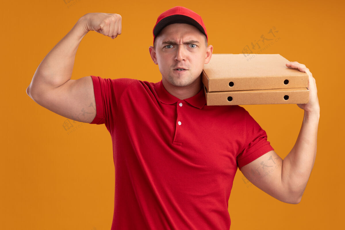 制服自信的年轻送货员身穿制服 肩上扛着披萨盒 在橙色的墙上显示出强烈的姿态帽子盒子肩膀