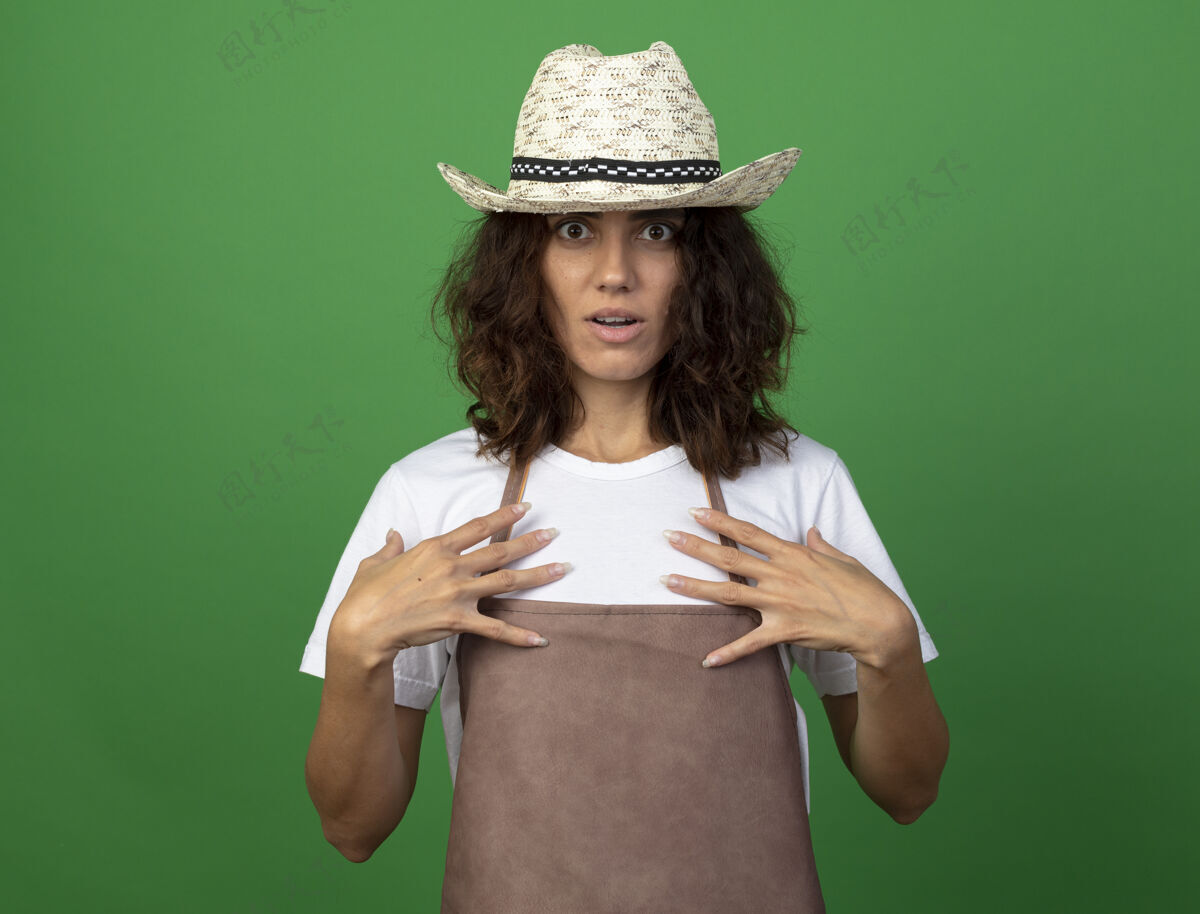 女人穿着制服的年轻女园丁惊讶地戴着园艺帽把手放在胸前胸部人女人