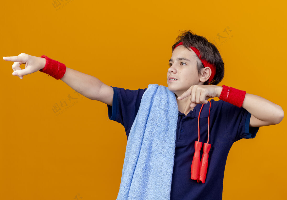 肩膀令人印象深刻的年轻英俊的运动男孩戴着头带和护腕与牙套和毛巾与跳绳在肩上看着并指着橙色墙上孤立的一面男孩牙齿人